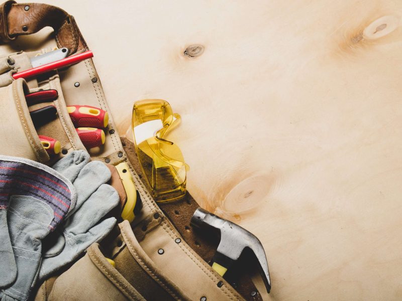 Tool belt with carpenter tools and protective wear on plywood sheet flat lay view