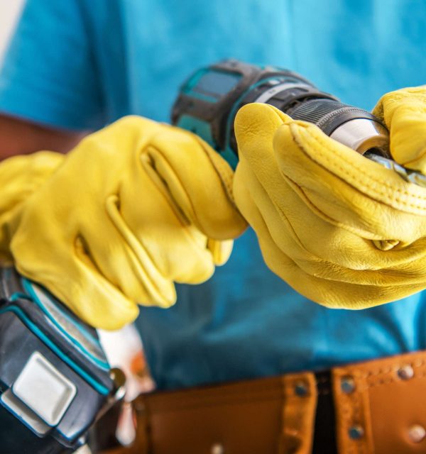 Closeup of Electrical Drill in Hands of Professional Construction Worker Wearing Yellow Protection Work Gloves. Professional Power Tools Theme.